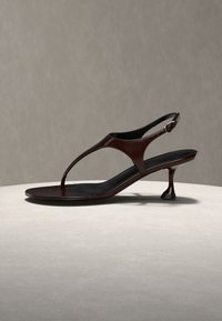 Ciemnobrązowa skórzana sandałka na obcasie z paskiem między palcami i regulowaną klamrą na neutralnej powierzchni i tle.