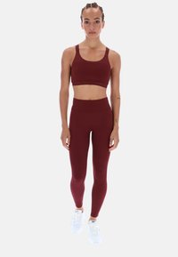 Burgundský sportovní podprsenka s širokými ramínky a logem, doplněná o vysoké burgundské legíny. Bílé sportovní boty dotvářejí celý vzhled.