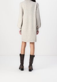 Femme debout de dos portant une robe en maille beige à manches longues et des bottes marron foncé à boucle arrivant au milieu du mollet sur un fond blanc.