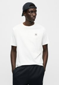 Λευκό βαμβακερό μπλουζάκι με στρογγυλή λαιμόκοψη και κοντά μανίκια, με μικρό στρογγυλό λογότυπο στο στήθος. Συνδυασμένο με μαύρο παντελόνι με ελαστική μέση.