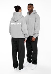 Felpa grigia con zip, con grande scritta bianca "MULTIPLY" sulla schiena, abbinata a pantaloni neri e scarpe da ginnastica bianche. Tessuto liscio, vestibilità rilassata.