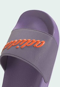 Fioletowe sandały na płaskiej podeszwie z teksturowanym, błyszczącym wykończeniem oraz szerokim paskiem z podniesionym pomarańczowym napisem. Gładka wewnętrzna powierzchnia.