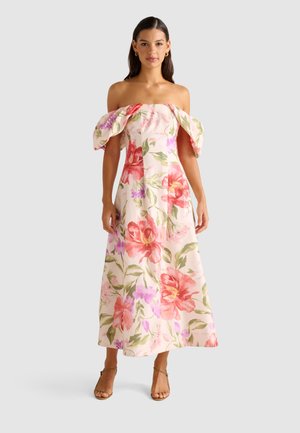 Una donna sta in modo elegante in un vestito floreale senza spalline con tonalità rosa e verdi, abbinato a sandali con cinturino, su uno sfondo neutro.