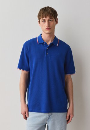 Ung mand står op ad en ensfarvet væg iført en blå poloshirt med rød og hvid kant og lyseblå jeans.