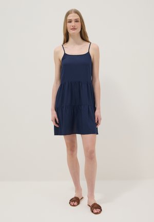 Jonge vrouw staat met een mouwloze navy blauwe jurk met verschillende lagen en bruine geweven platte sandalen tegen een eenvoudige lichte achtergrond.