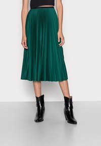 Vihreä laskostettu midi-hame, jossa on teksturoitu pinta, käytössä mustien nilkkasaappaiden kanssa. Vyötäröllä on hienovarainen logoyksityiskohta.