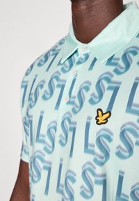 Polo vert menthe avec un motif abstrait répété et un petit logo d'oiseau jaune sur la zone gauche de la poitrine. Col à boutons.