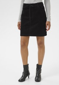 Jupe en velours noir avec une coupe au genou, des poches avant et une ceinture à nouer. Associée à des bottines noires et un haut à manches longues gris clair.