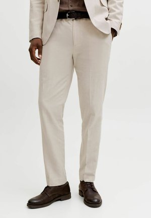 Man die beige pantalon draagt, een donkerbruine leren riem en donkerbruine nette schoenen, staand met ontspannen handen, tegen een witte achtergrond.