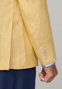Blazer en lin jaune avec une finition texturée, comprenant quatre boutons beiges au niveau des poignets et une chemise rayée bleue et blanche en dessous.
