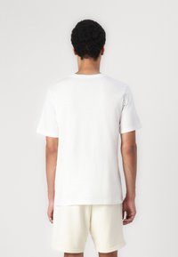 Weißes Baumwoll-T-Shirt mit Rundhalsausschnitt und kurzen Ärmeln. Glatte Textur, lockere Passform, von hinten betrachtet. Kombiniert mit hellen Shorts.