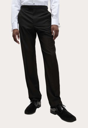 Pantalones formales negros con un corte ajustado, textura suave y un ligero brillo. Combinados con zapatos negros brillantes que exhiben un diseño clásico.
