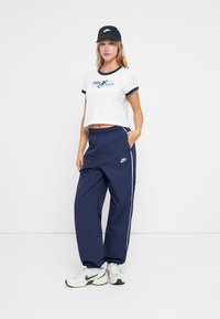 Balta īsā t-kreka ar tumši zilām apmalēm un Nike logo; tumši zilas sporta bikses ar baltām sānu svītrām un Nike logo; melna cepure un sporta apavi.