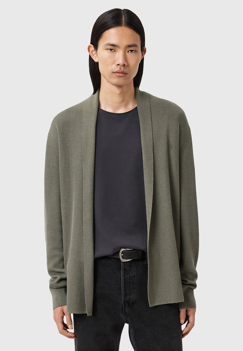Cardigan ouvert, de couleur olive, en tricot côtelé texturé, avec des manches longues et une coupe décontractée, porté sur un T-shirt sombre et un jean noir.