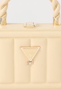 Bézs műbőr kézitáska steppelt mintával, csavart felső fogantyúval, aranyszínű fém részekkel és háromszög alakú logóval, amelyen a "GUESS" és a "girl" felirat szerepel.