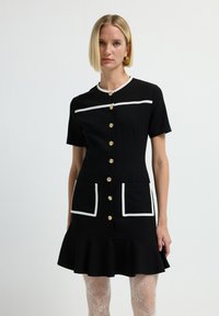 Vestito nero a maniche corte con rifiniture bianche, chiusura con bottoni, due tasche frontali e orlo svasato. Bottoni dorati aggiungono un contrasto.