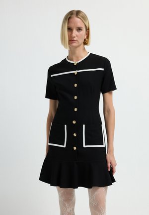 Vestito nero a maniche corte con rifiniture bianche, chiusura con bottoni, due tasche frontali e orlo svasato. Bottoni dorati aggiungono un contrasto.