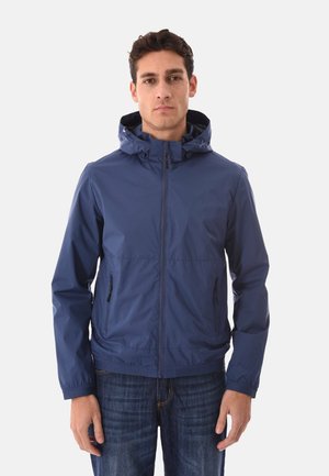 Uomo che indossa una giacca con cappuccio blu navy con cerniera e tasche laterali, jeans blu scuro, in piedi contro uno sfondo chiaro e uniforme.