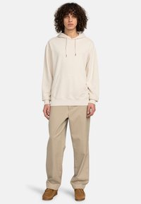 Sudadera beige clara con un bolsillo frontal, capucha con cordón, y un corte relajado, combinada con pantalones holgados beige y calzado marrón claro.