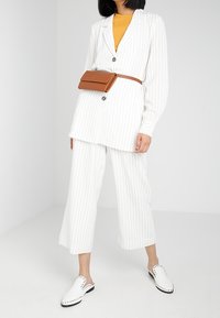 Valkoinen raidallinen bleiseri napilla, yhdistettynä sinapinkeltaiseen puseroon ja valkoisiin capri-housuihin. Ruskea vyölaukku ja valkoiset läpsyt, joissa on niittejä.