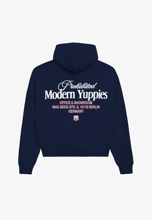 Młoda osoba ubrana w białą czapkę, czarną bluzę z kapturem, krótkie spodenki moro, białe skarpetki i brązowe buty, stoi na jednolitym tle.