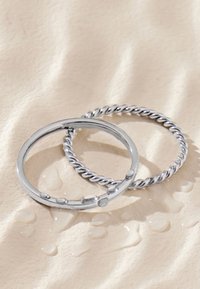 Zwei silberne Ringe auf einer beigen, strukturierten Oberfläche mit Wassertropfen; ein Ring ist glatt mit kleinen erhabenen Punkten, der andere ist im gedrehten Seil-Stil.