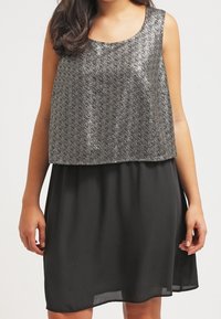 Šaty bez rukávů s lesklým kovov�ým stříbrným vrškem a splývavou černou sukní. Hladký textilní materiál s uvolněným střihem.