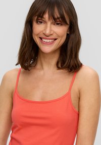Koronkowy top w kolorze koralu wykonany z miękkiej, elastycznej tkaniny. Posiada cienkie ramiączka oraz okrągły dekolt, charakteryzuje się gładką fakturą i dopasowanym krojem.