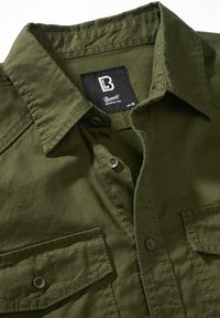 Oljčno zelena srajca na gumbe z ovratnikom, dvema sprednjima žepoma in šivanskimi detajli. Material se zdi trpežen in teksturiran. Vidna je znamka.