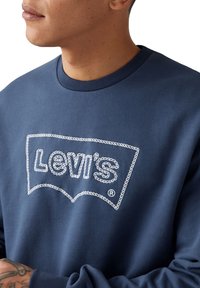 Marineblauwe sweatshirt met witte geborduurde "Levi's" logo in een afgerond embleemontwerp. Zachte textuur met geribbelde halslijn en boorden.