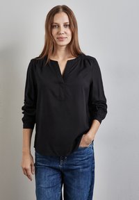 Blouse noire à manches longues avec un col en V et des épaules froncées, fabriquée dans un tissu lisse. Associée à un jean en denim bleu.