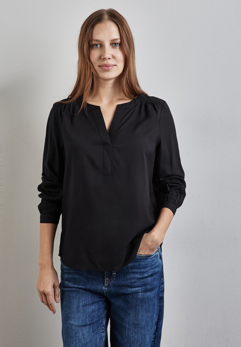 Blouse noire à manches longues avec un col en V et des épaules froncées, fabriquée dans un tissu lisse. Associée à un jean en denim bleu.