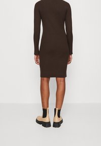 Personne portant une robe marron côtelée moulante à manches longues, assortie à des bottines beiges à semelle épaisse, sur un fond uni.