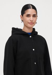 Vrouw met donker haar dat naar achteren is getrokken, die zilveren bloemvormige oorbellen draagt en een zwarte gewatteerde jas met zilveren drukknoopsluitingen.