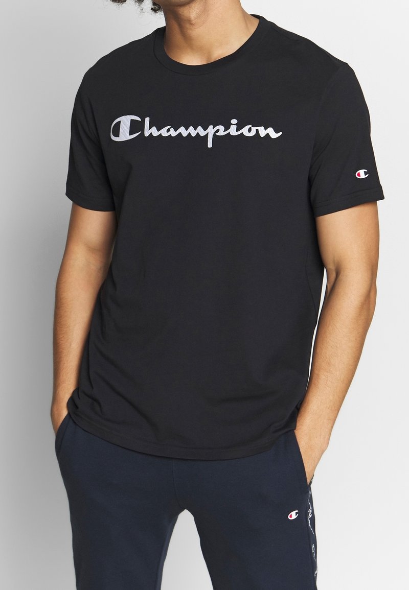 Muž v černém tričku Champion a námořnických modrých teplákách, s rukama v kapsách, s krátkým střihem na krku a u poloviny stehna.