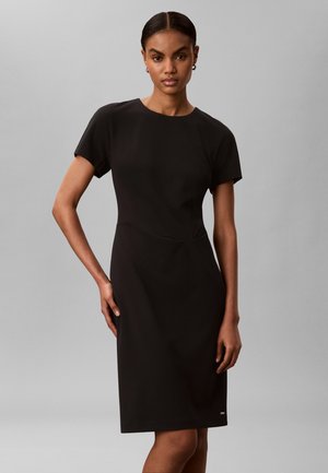 Γυναίκα στέκεται με το χέρι στη μέση φορώντας μαύρο φόρεμα με κοντά μανίκια μέχρι το γόνατο, σε απλό γκρι φόντο.