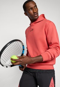 Sweatshirt à capuche corail avec logo noir, associé à un pantalon de sport noir. Tient une balle de tennis verte et une raquette de tennis.