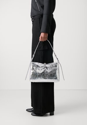 Sac à main métallique argenté avec une texture froissée, de forme rectangulaire plate, une courte poignée et une fermeture à clapet, tenu par une personne en tenue noire.