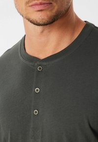 Maglietta henley in cotone grigio scuro con colletto rotondo, patta con tre bottoni e dettagli in metallo, e una cucitura a forma di X sul bordo.