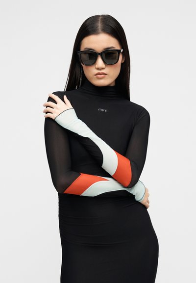 Γυναίκα με μακριά σκούρα μαλλιά, που φορά μεγάλα μαύρα γυαλιά ηλίου και μαύρο φόρεμα με ανοιχτογάλανα και κόκκινα σατινέ μανίκια, σταυρώνει τα χέρια της.