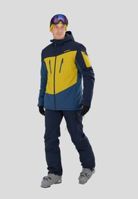 Herreskibeklædning med en farvet blok-jakke i navy og gul, med lynlåsede lommer; parret med navy bukser og lysegrå skistøvler.