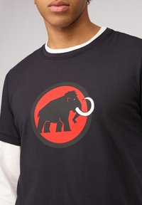 Zwarte T-shirt met korte mouwen met een rood circulair logo met een zwart mammoth graphic en een witte slagtanddetail. Onderlaag met een witte kraag.