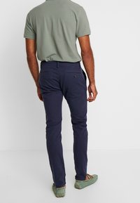 Pantaloni navy in cotone con vestibilità slim, dotati di tasche posteriori con chiusura a bottone e una texture liscia. Indossati con una camicia verde e scarpe.