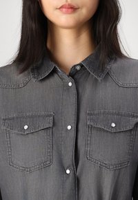 Chemise en denim grise avec un col classique, des boutons-pression et deux poches poitrine avec rabats. Le tissu a une texture lisse avec des coutures légères.