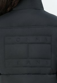Black padded jacket with a stitched logo panel showing "TOMMY JEANS" on the back. Smooth texture and minimalistic design.