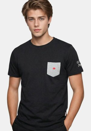 Jeune homme aux cheveux châtain clair portant un t-shirt noir avec une poche grise ornée d'un petit losange rouge.