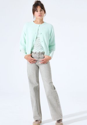 Minzgrüner Cardigan mit runden Knöpfen über einem Spitzentop. Graue Hosen mit weitem Bein und dezenten Punktmustern. Modell steht mit Händen in der Hüfte.