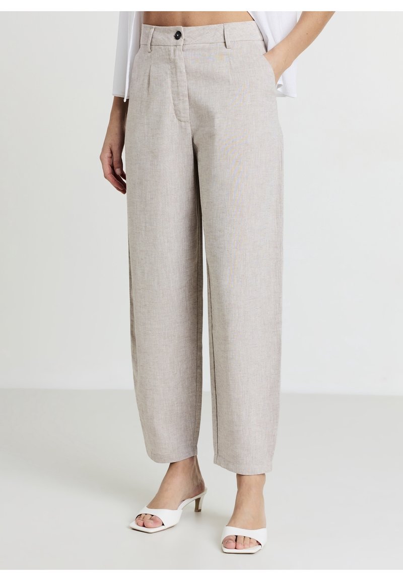 Pantalon beige à jambes larges avec bouton à l’avant et poches, associé à des sandales blanches à talons ouverts, porté par une personne debout.