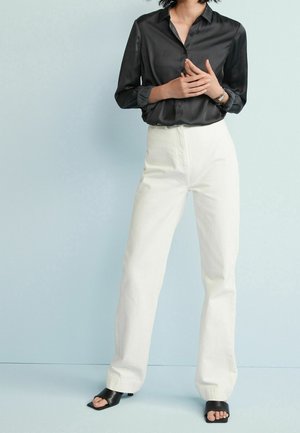 Black satin button-up shirt, white high-waisted straight-leg pants, and black slide sandals, set against a light blue background.