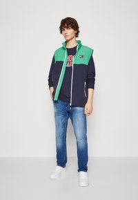 Chaleco color bloque en verde y marino, con cierre de cremallera, y una camiseta de cuello redondo en marino por debajo. Combinado con jeans de un azul claro y zapatillas blancas.